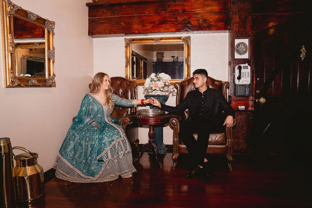 A couple in vintage attire sit facing each other in an elegantly decorated Victorian-style room with antique furnishings.
