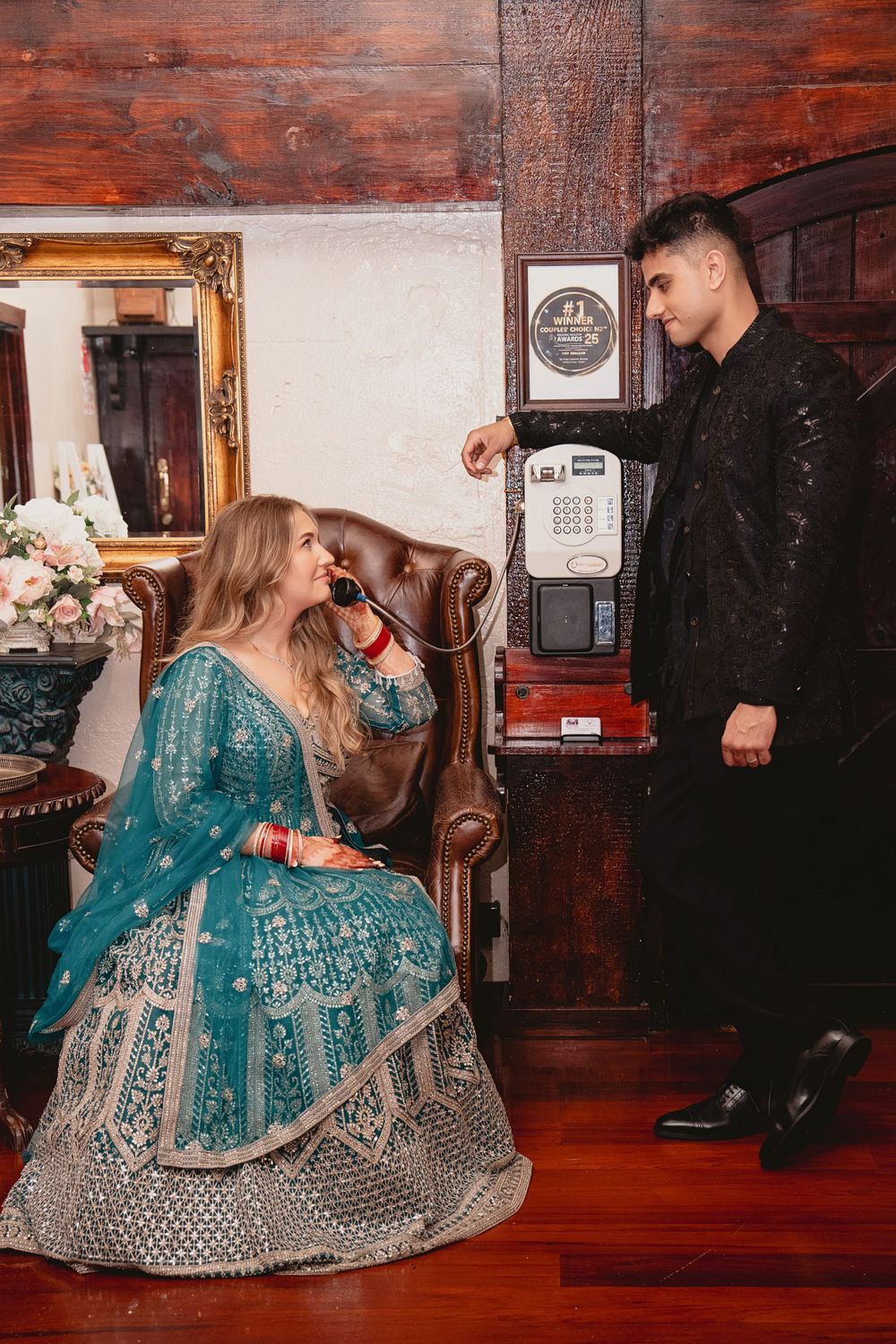 Bride in teal lehenga sits in vintage chair using retro phone while groom in black sherwani stands beside her.