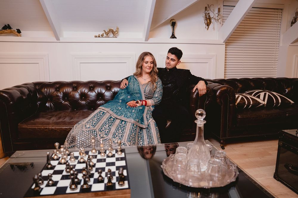 Couple in elegant Indian attire posing beside a chess board in a luxurious room with leather sofa.