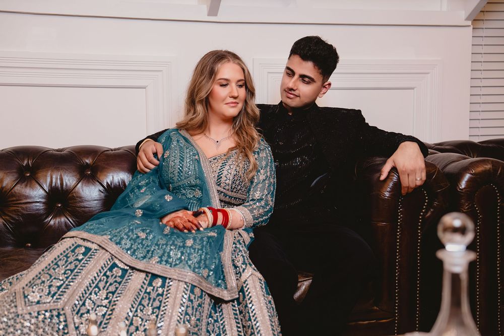 Couple in elegant Indian attire sitting on leather sofa, woman in teal lehenga, man in black suit.