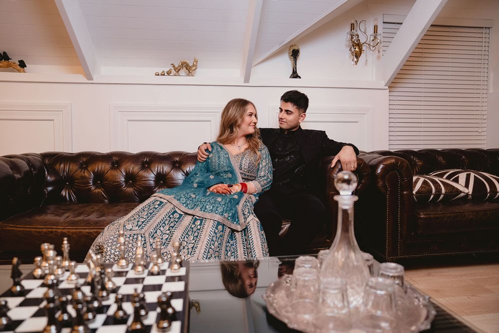 Couple in elegant Indian attire sitting together on a leather sofa beside a chess set and crystal decanter.