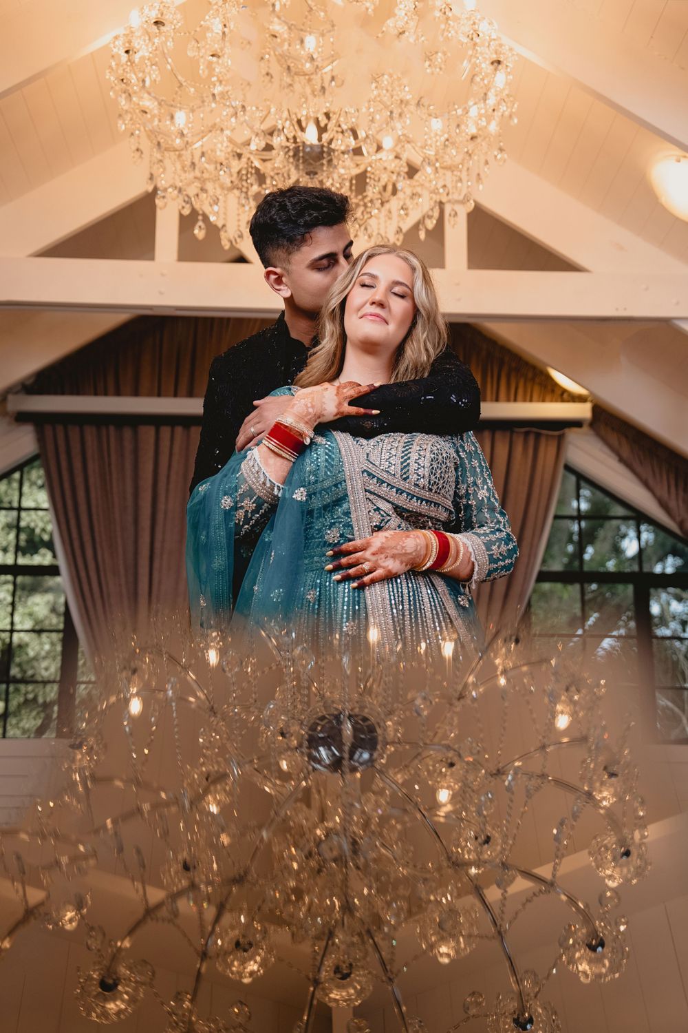 Couple in Indian wedding attire embrace under crystal chandelier, bride in teal lehenga with mehndi, groom in black sherwani.