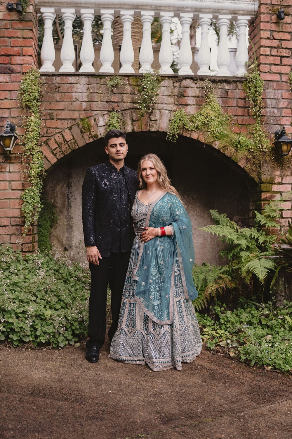 Indian wedding couple poses before brick arch, bride in teal lehenga and groom in black sherwani outdoors.