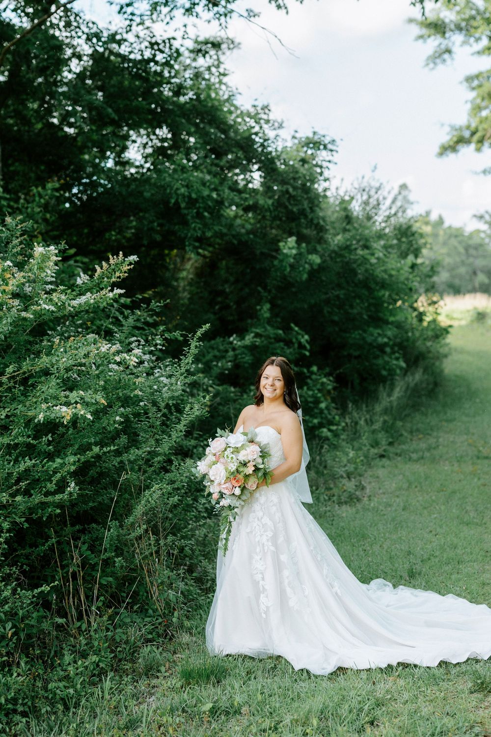 Bride in strapless lace A-line gown holds pink bouquet while posing in lush green garden setting.