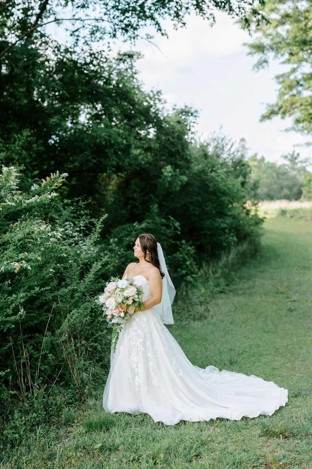 Bride in lace gown and veil holds floral bouquet outdoors near lush green trees on her wedding day.