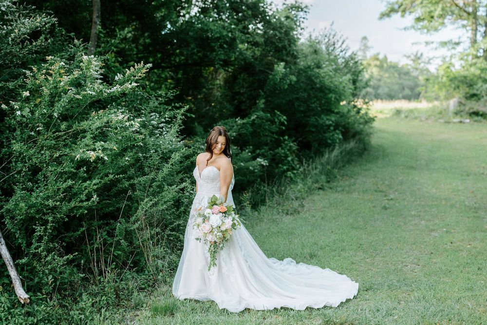 Bride in white gown holding bouquet stands on green lawn beside lush trees in outdoor wedding portrait.