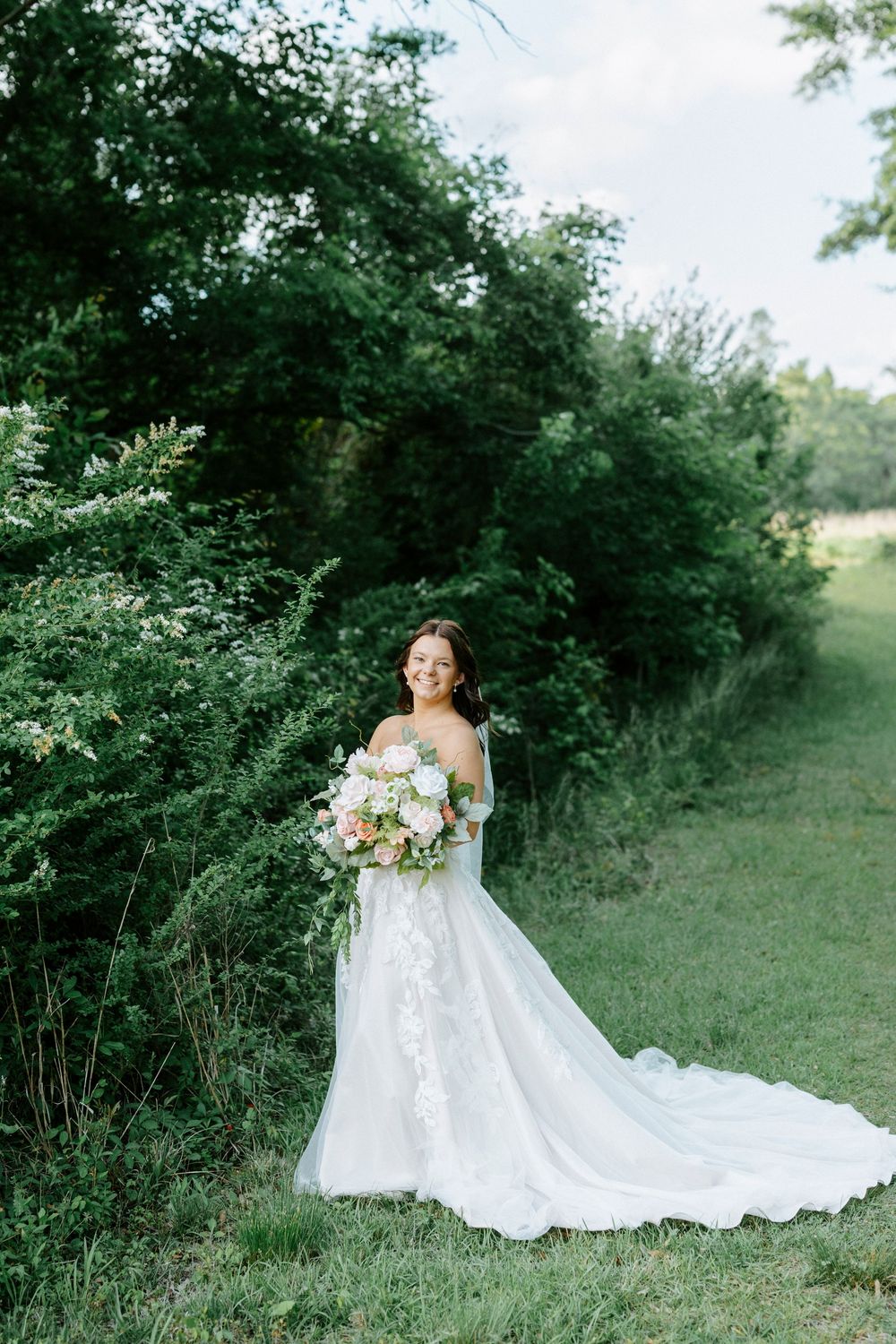 Smiling bride in off-shoulder lace wedding gown holding pink floral bouquet, standing in lush green garden setting.