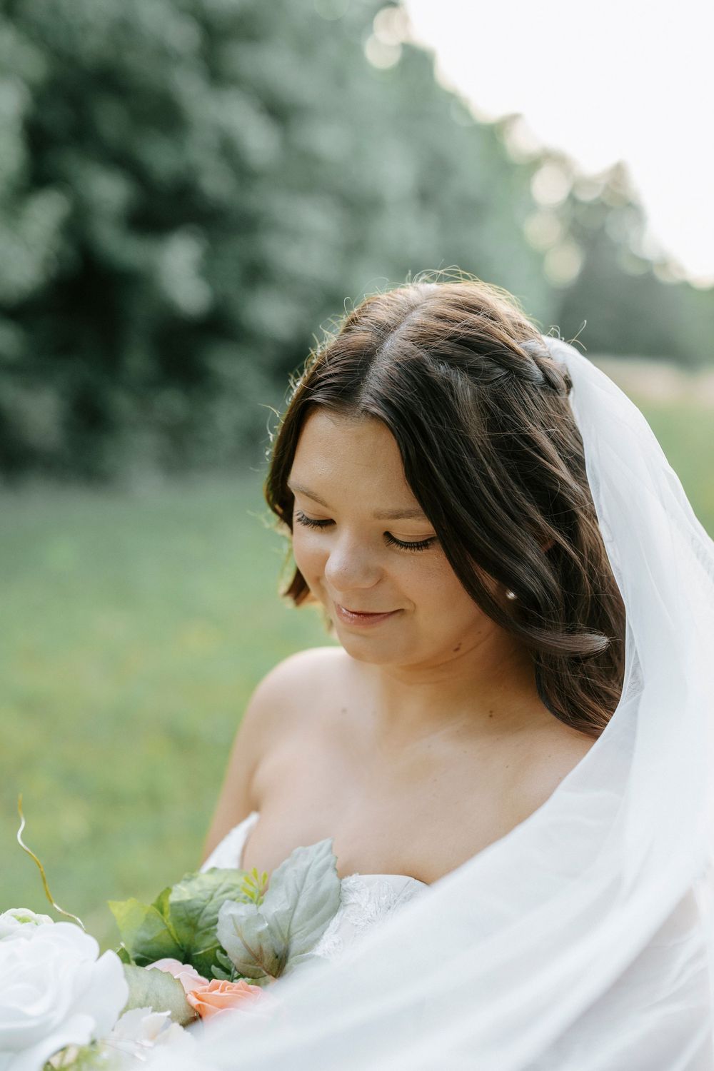 A bride in a white gown and veil smiles softly while holding a bouquet of white and peach flowers outdoors.