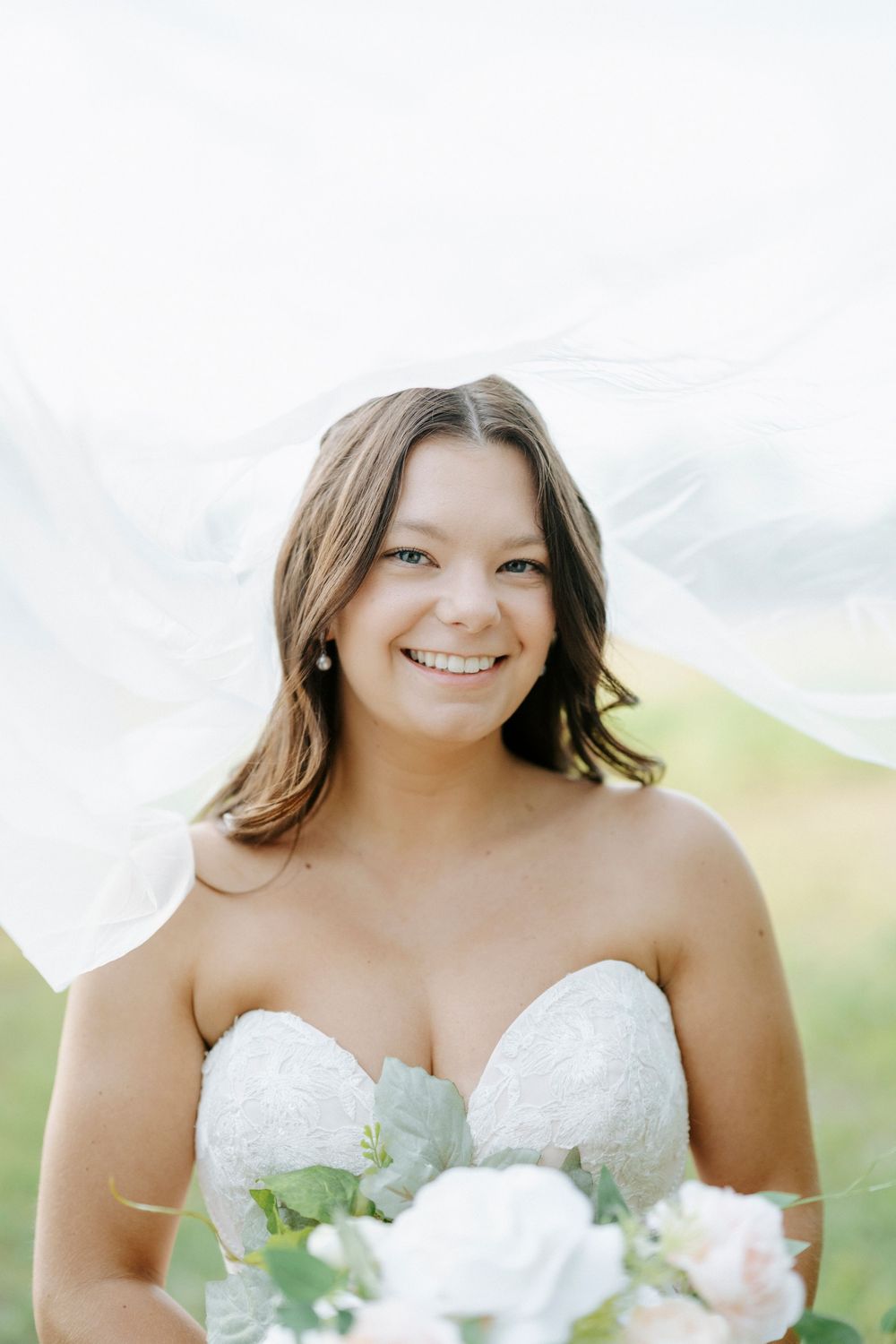 Smiling bride in strapless lace gown holds white bouquet under flowing veil outdoors.