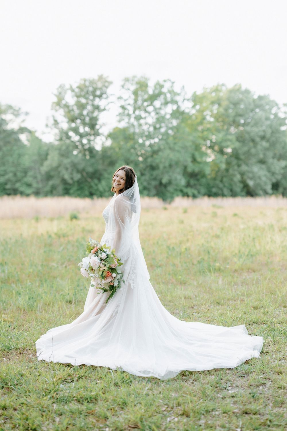 Bride in white gown with long train and veil holds bouquet in green field with trees.