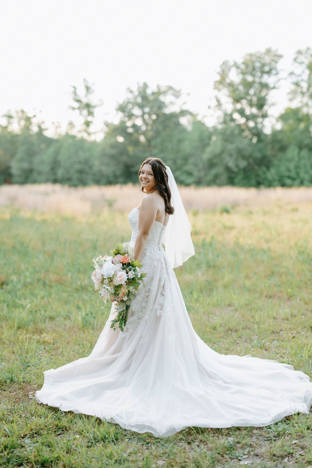 Smiling bride in lace wedding gown with long train and veil holds bouquet in sunlit meadow.