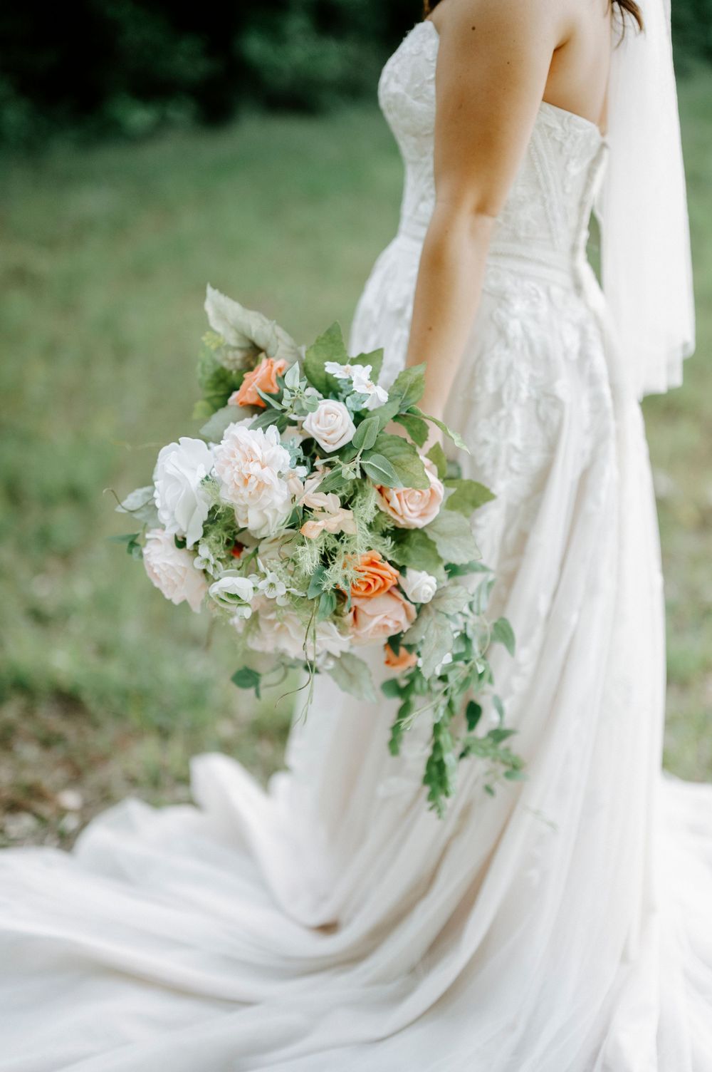 Bride in a strapless lace wedding gown holding a cascading bouquet of peach and white roses with greenery.