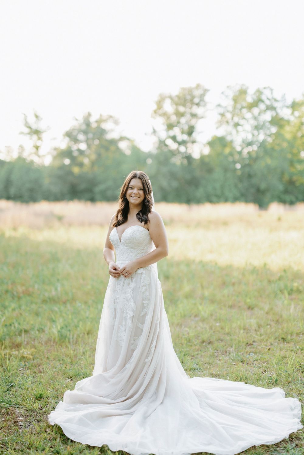 Smiling bride in a white lace wedding gown with a long train standing in a sunlit grassy field.