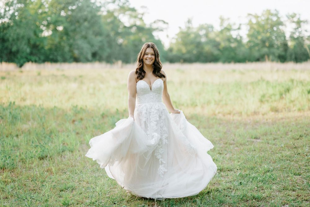 Bride twirling in a white lace wedding gown in a sunlit green field, smiling joyfully outdoors.
