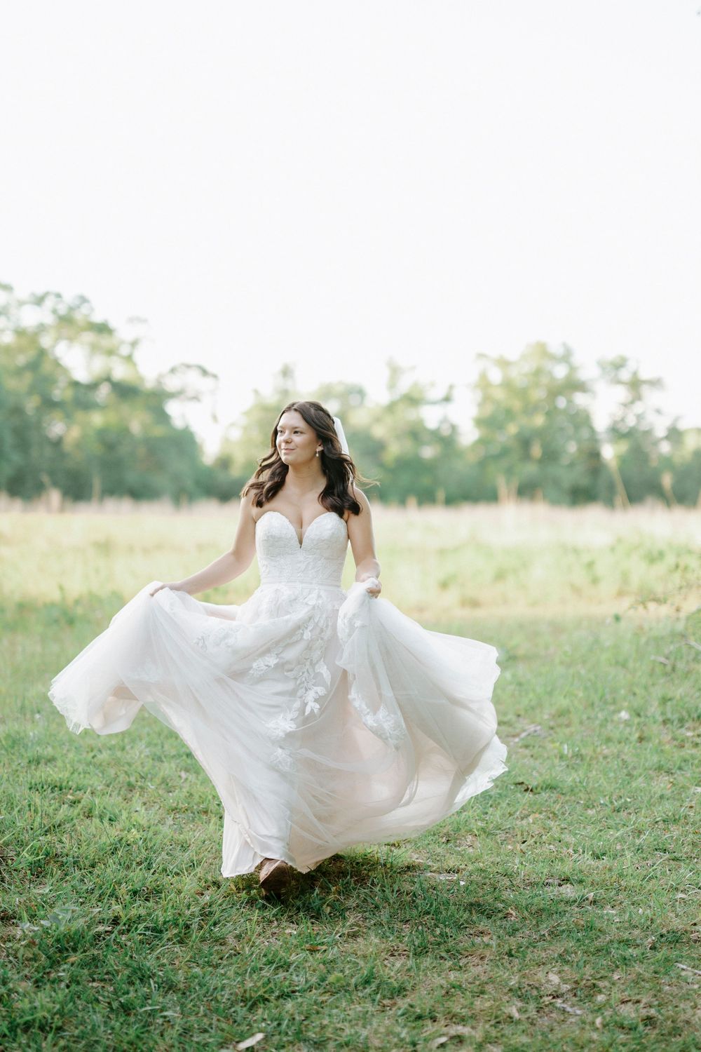 Bride in flowing white lace gown twirling in a green field, with trees in the background on her wedding day.