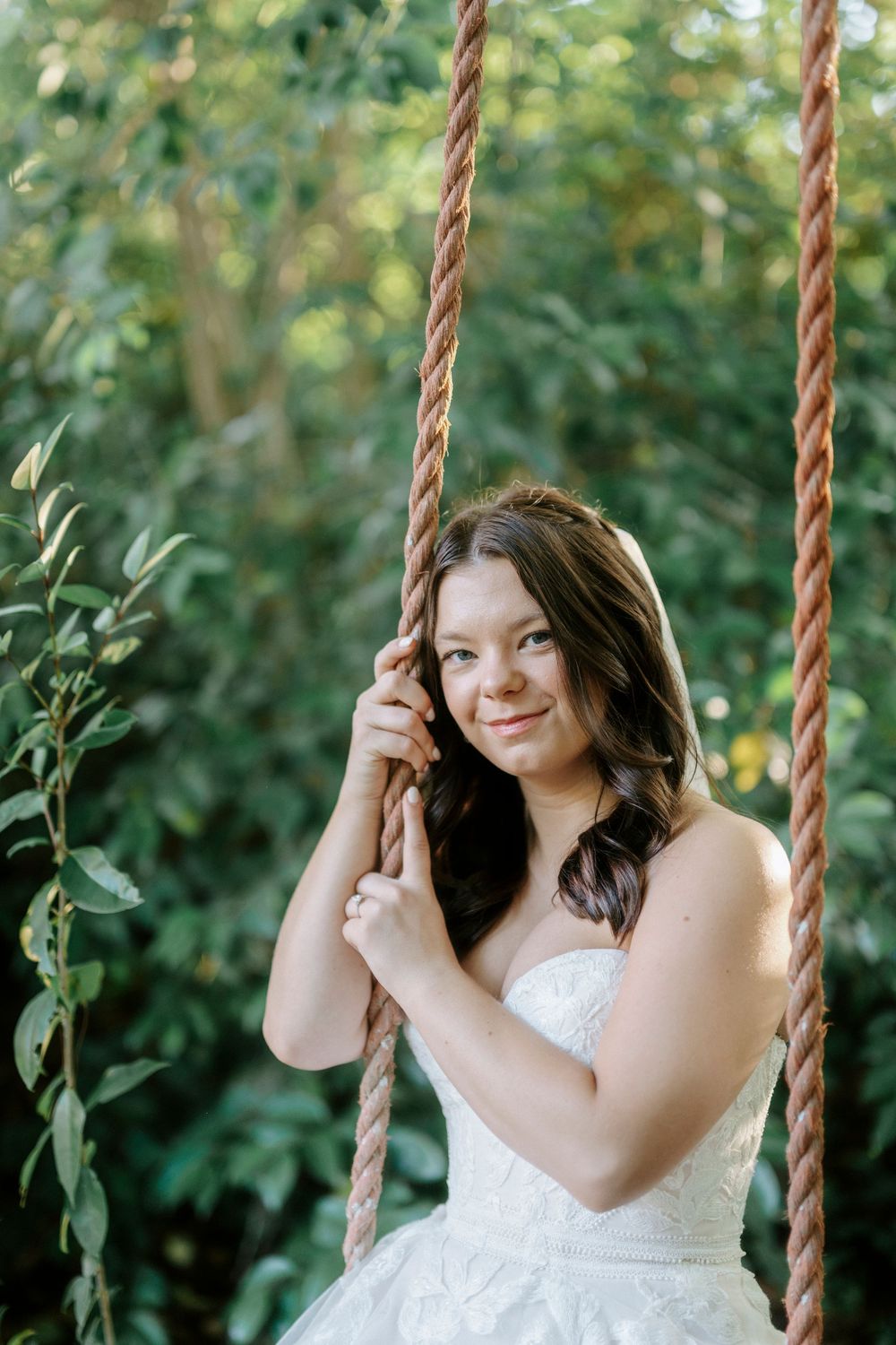Bride in white strapless gown sitting on a rope swing surrounded by lush green foliage, smiling softly.