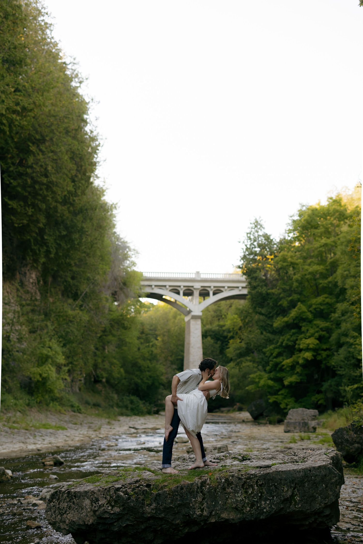Couple sharing a romantic dip kiss on rocks below an arched bridge surrounded by lush green trees.