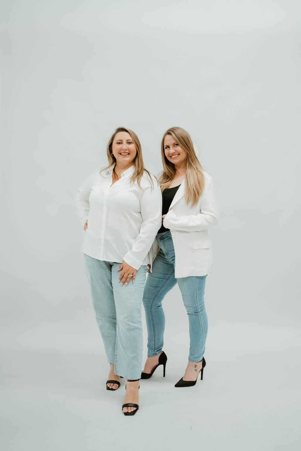 Two smiling women in white tops and jeans posing together against a light gray background.