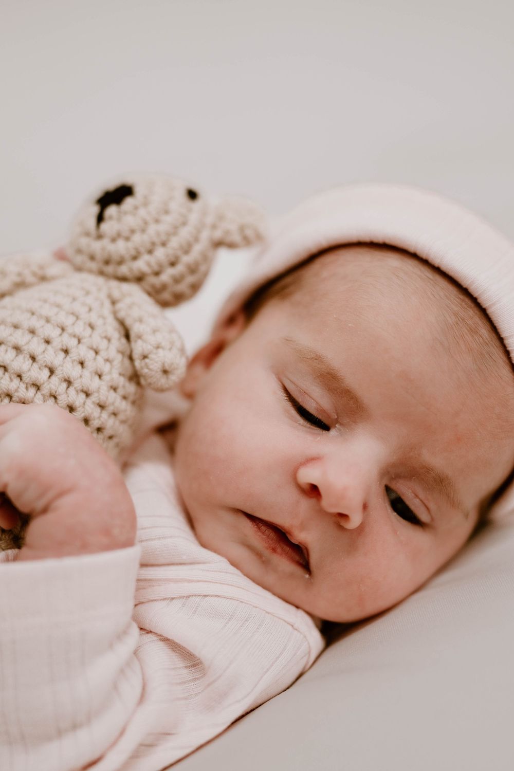 Sleeping newborn baby in pink outfit cuddles a crocheted teddy bear toy.