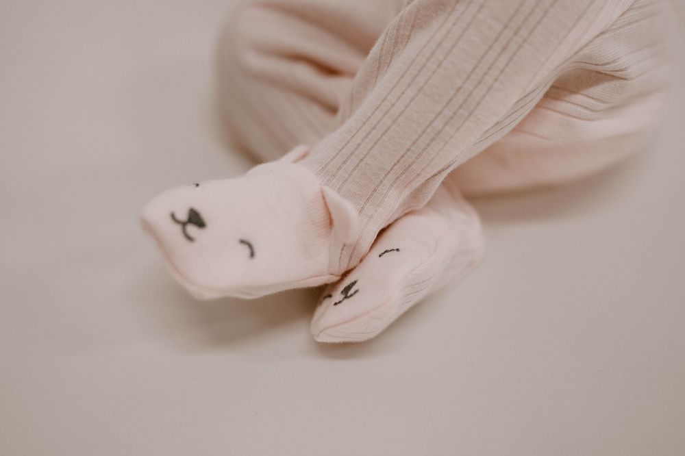 Close-up of baby feet wearing cute animal-face booties with a soft pink ribbed onesie on a neutral background.