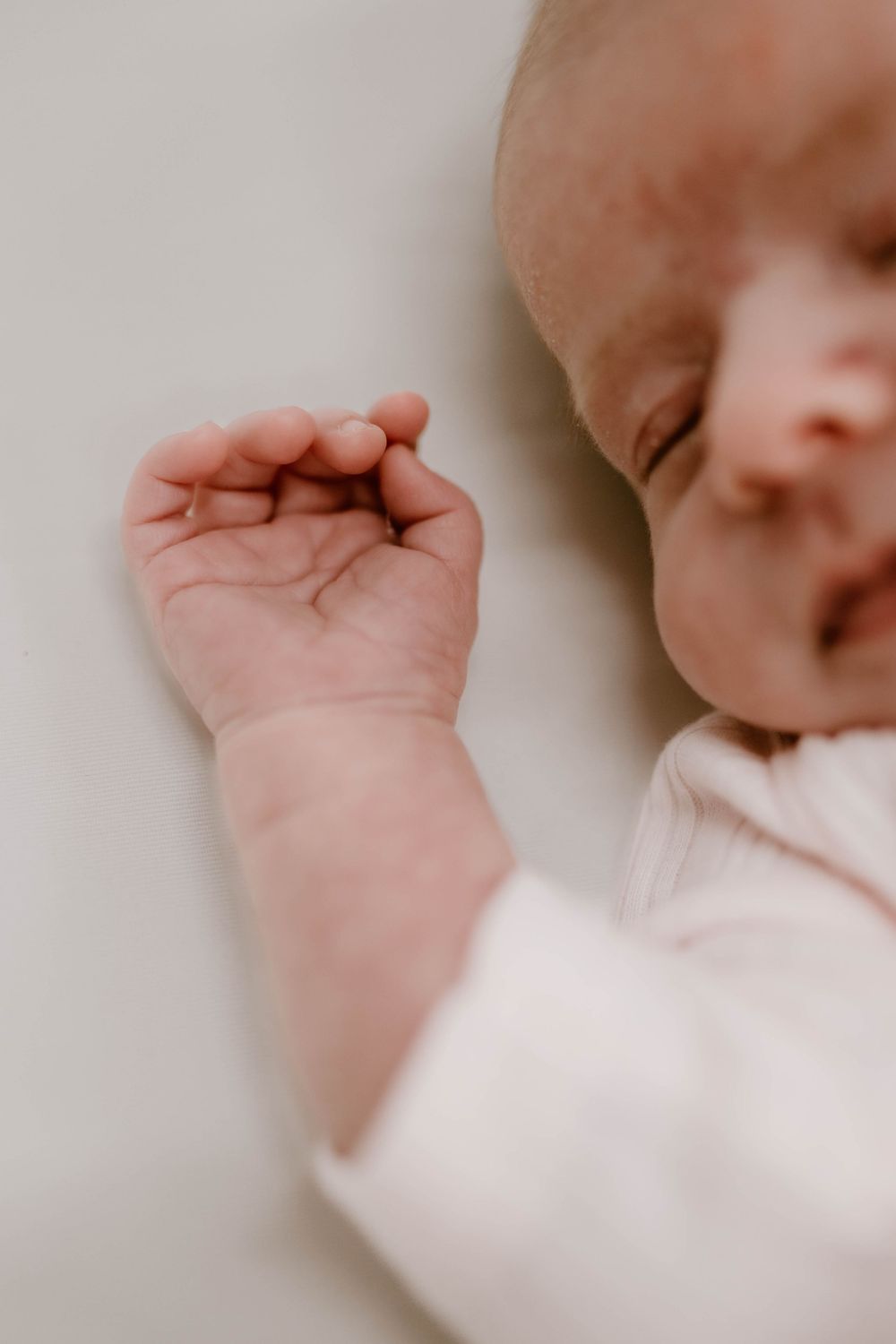 Close-up of a sleeping newborn baby's tiny hand and peaceful face resting on a white surface.