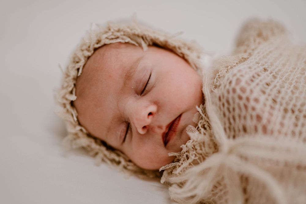 Sleeping newborn baby wrapped in a soft beige knit blanket and matching bonnet on a white background.