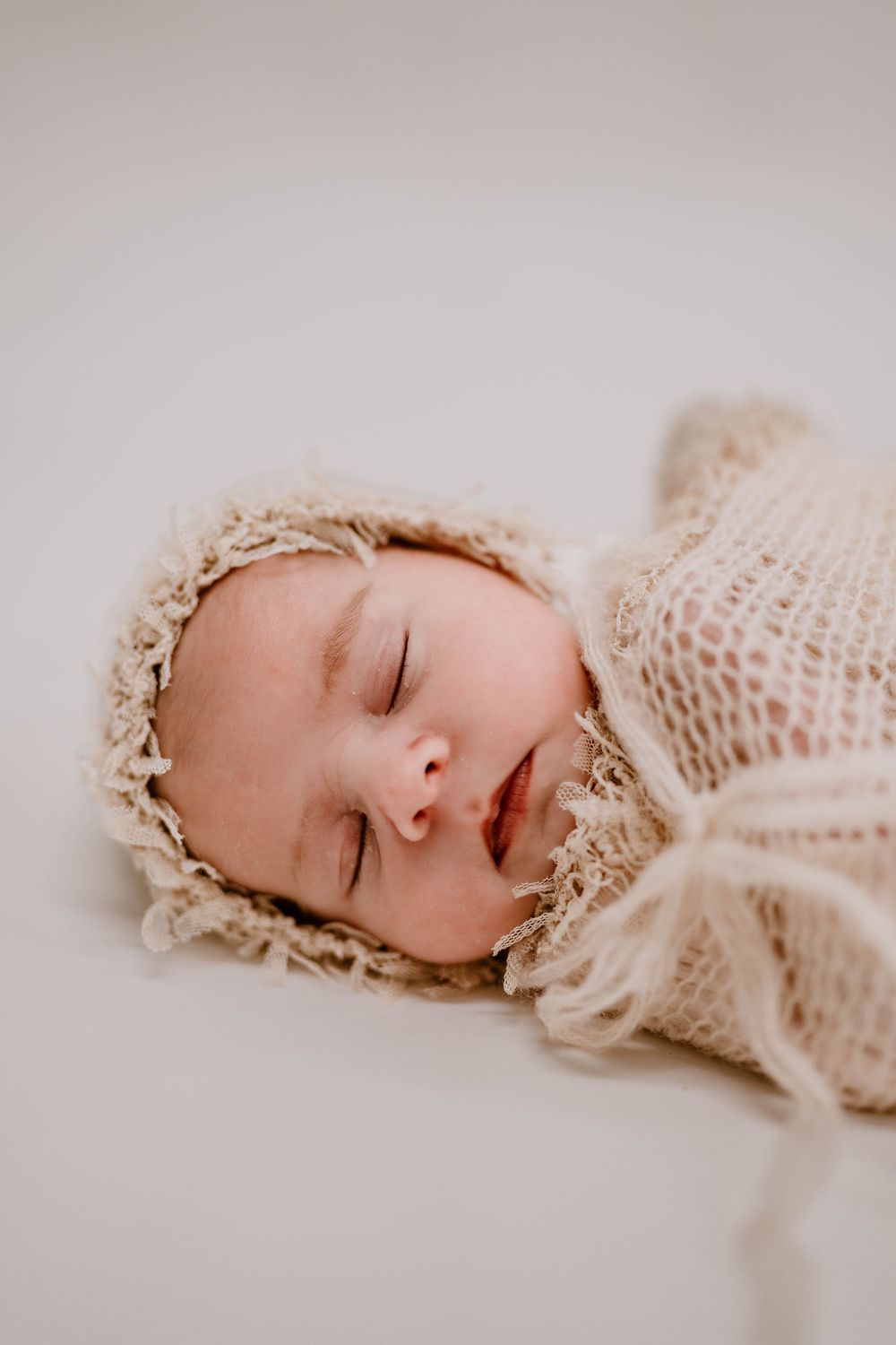 Sleeping newborn baby wrapped in a cream knit lace swaddle and bonnet on a white background.