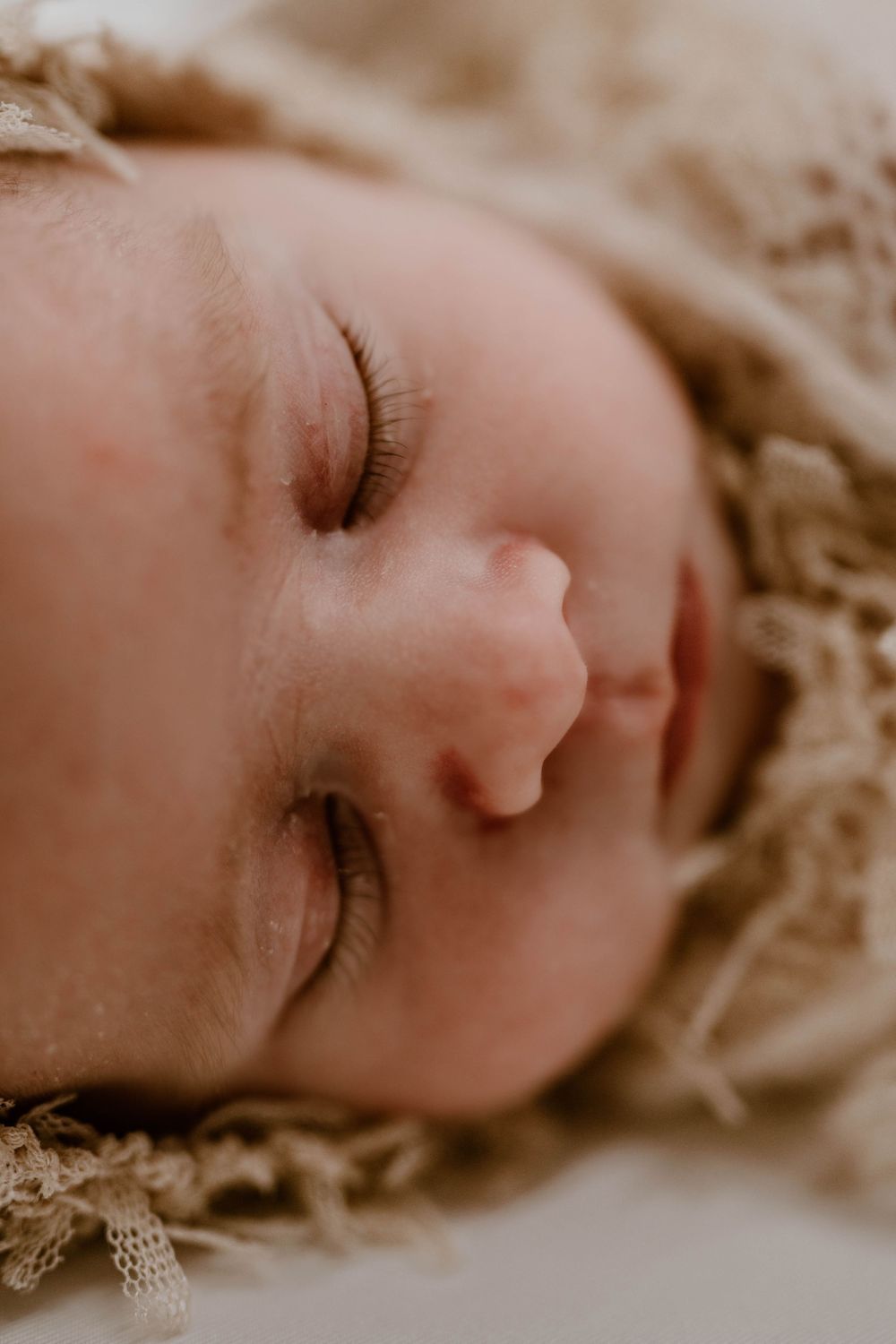 Macro close-up of a serene sleeping newborn baby's face with delicate eyelashes on a soft beige wrap.