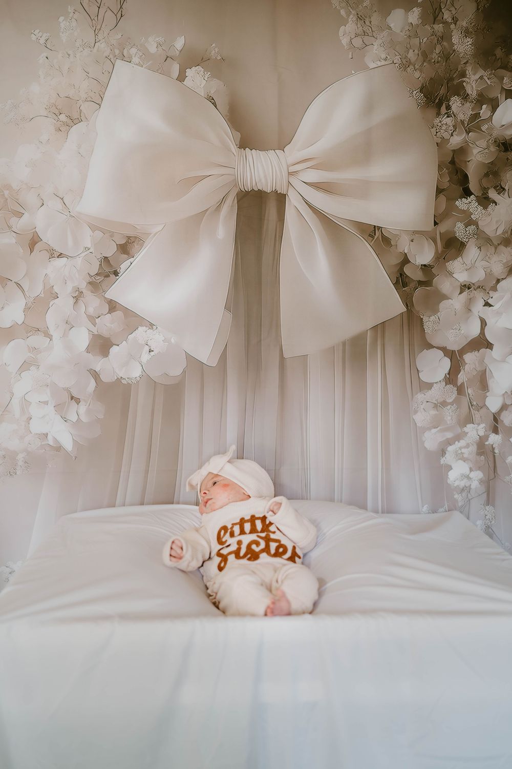 Newborn baby in little sister outfit lies under elegant white bow canopy.