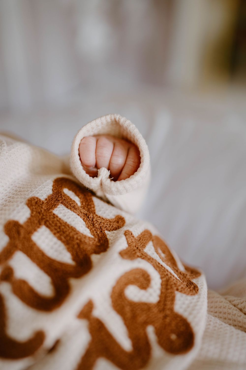 Close-up of a newborn baby's tiny fist wrapped in a cream knit mitten, resting on a personalized embroidered blanket.