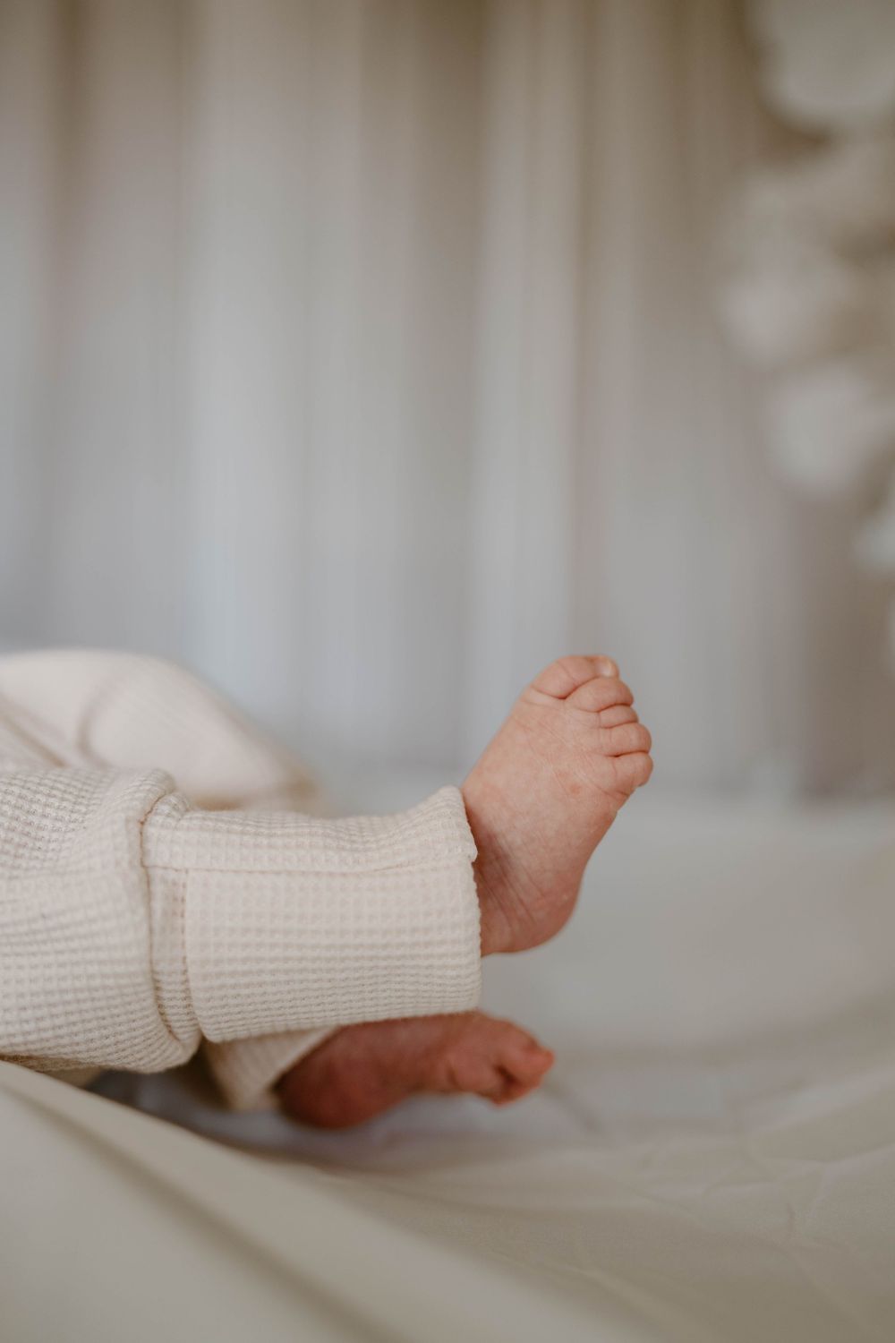 Close-up of tiny newborn baby feet in a white waffle knit onesie resting on a soft white surface.