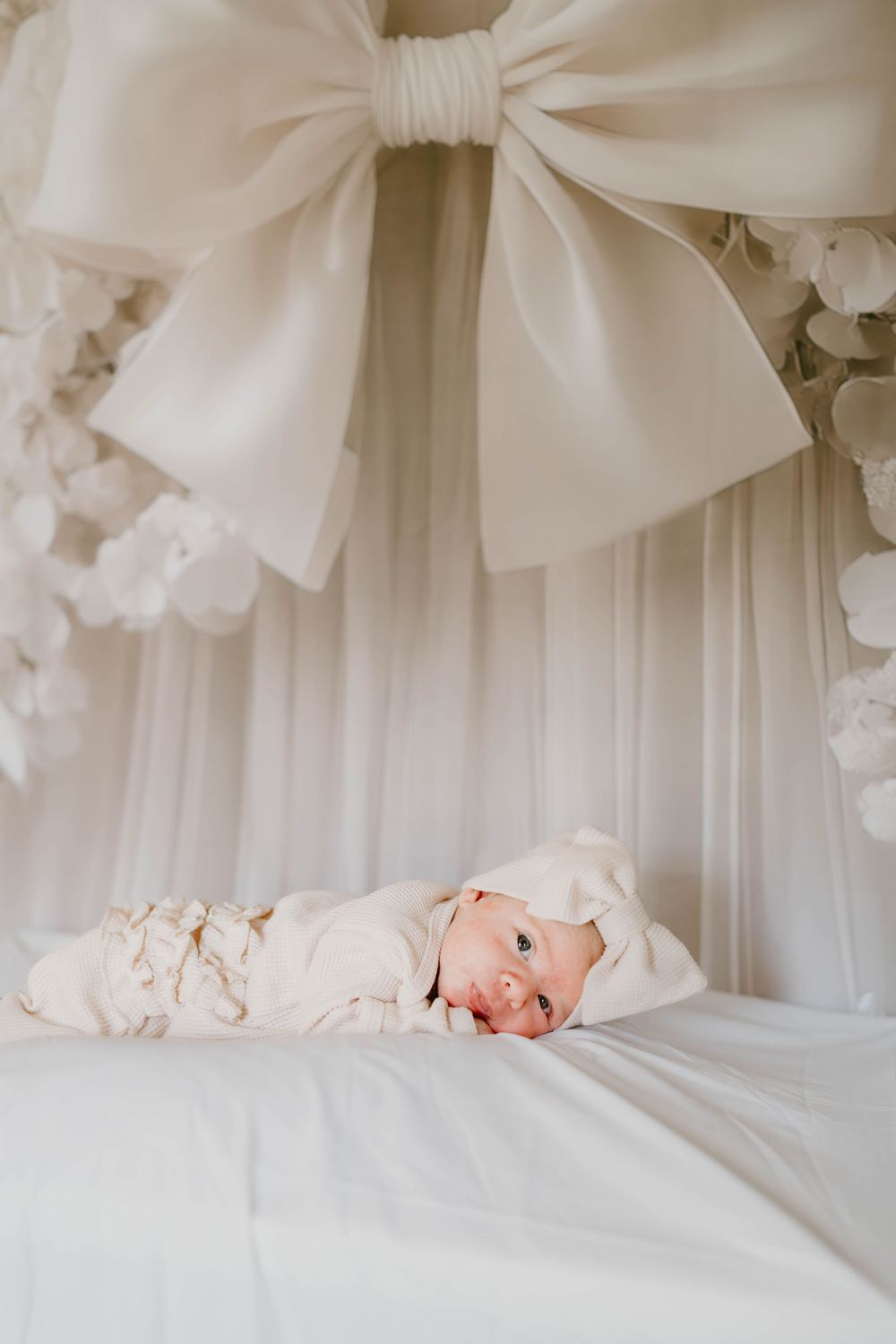 Peaceful newborn baby in white outfit rests under decorative bow canopy.