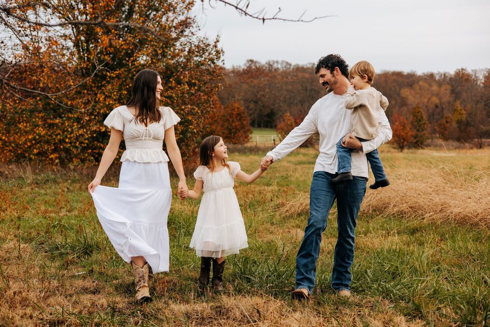 A happy family walks together in a fall meadow, parents holding hands with two young children.