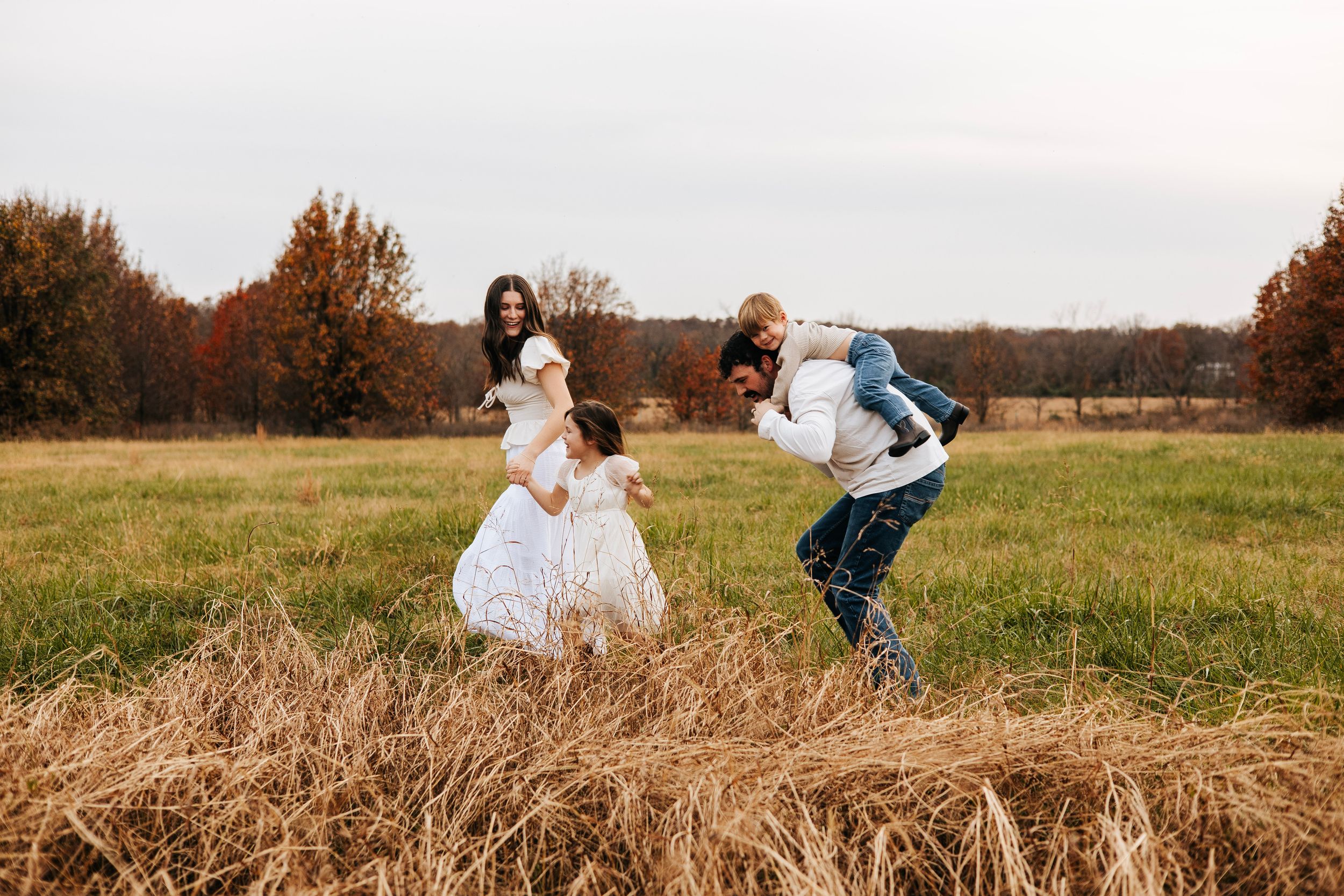 Family playing in an autumn field, parents lifting children near golden hay bales with colorful fall trees in background.