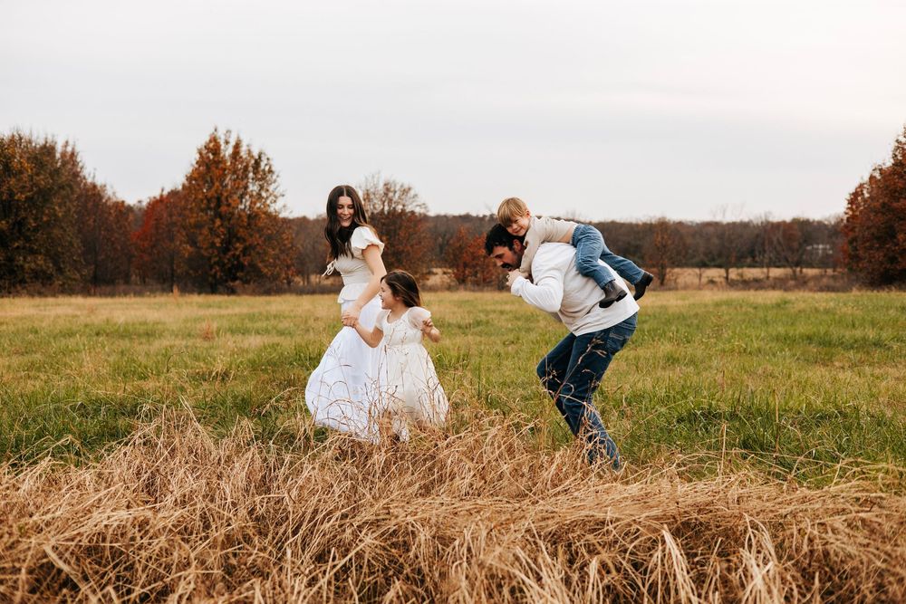 Family playing in an autumn field, parents lifting children near golden hay bales with colorful fall trees in background.