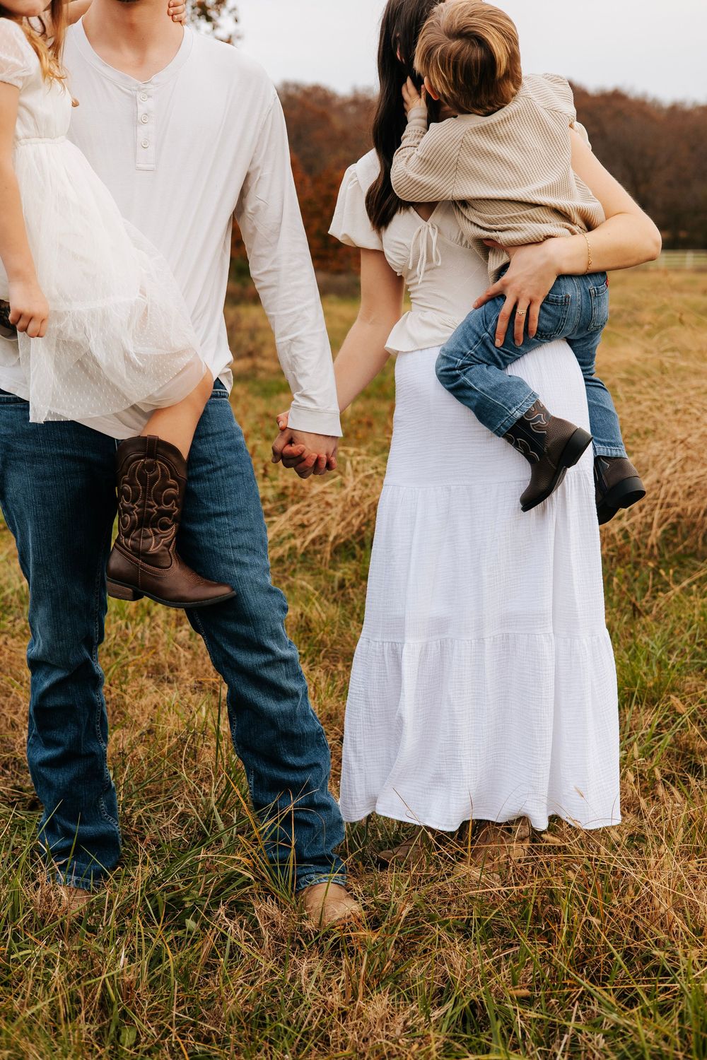 Family in white tops and jeans walking through autumn field, mother in white maxi dress holding toddler.
