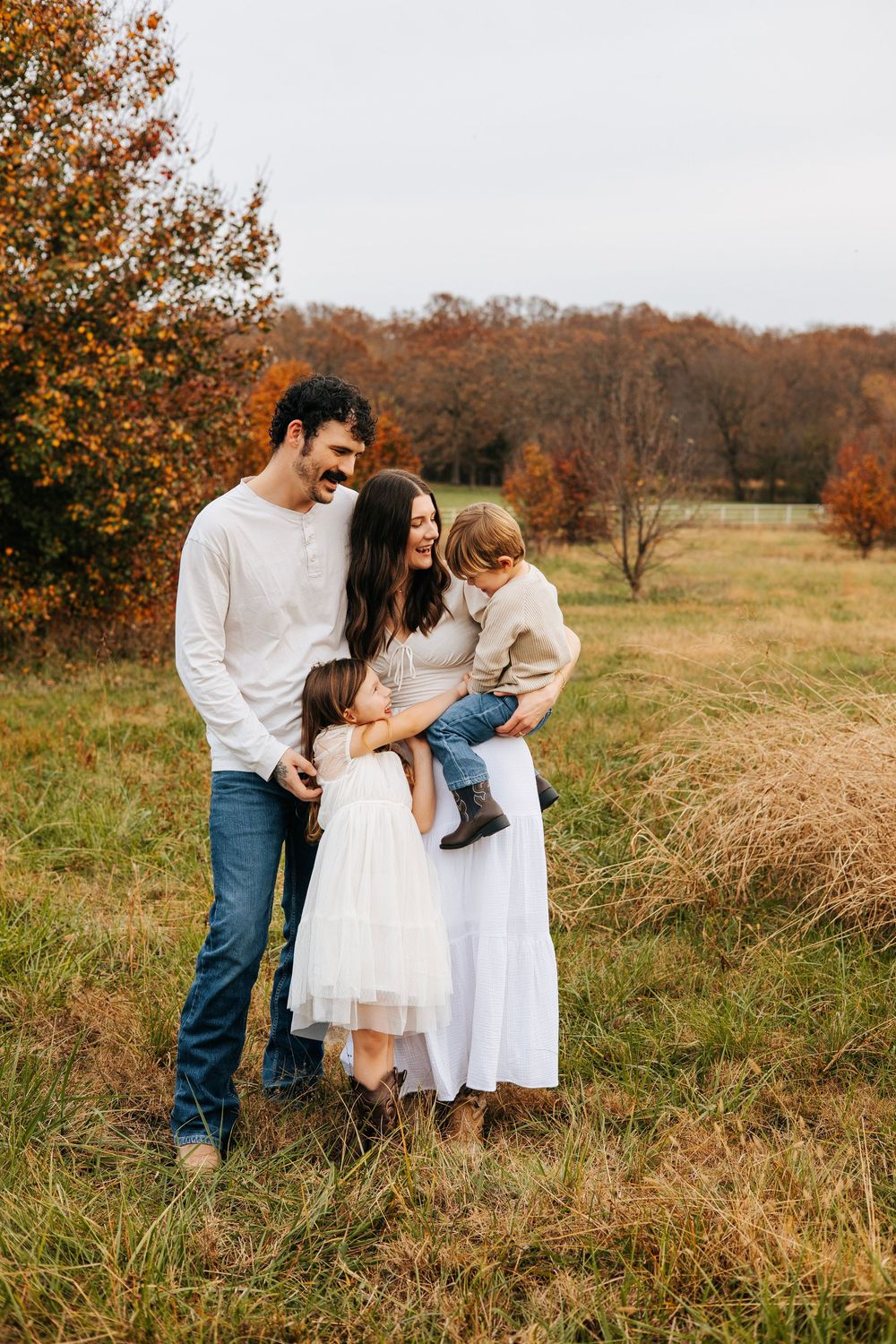Family of four poses in autumn field, parents smiling at toddler son held by mom in white dress.