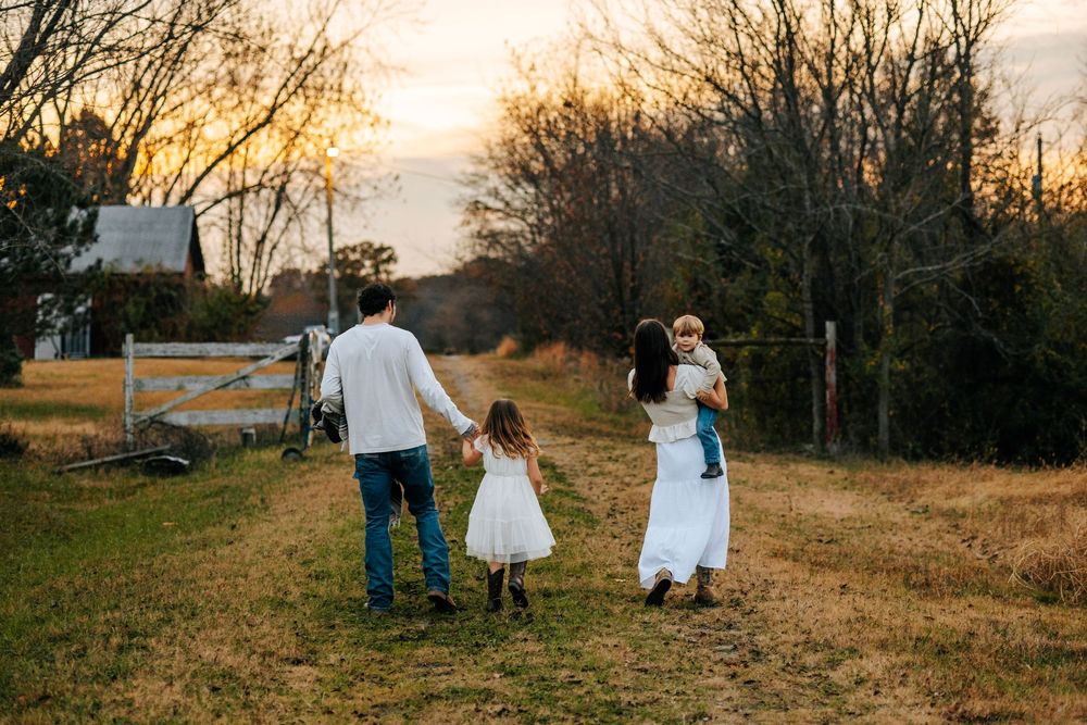 Family of four walking toward sunset on rustic farm path, children in white dresses.