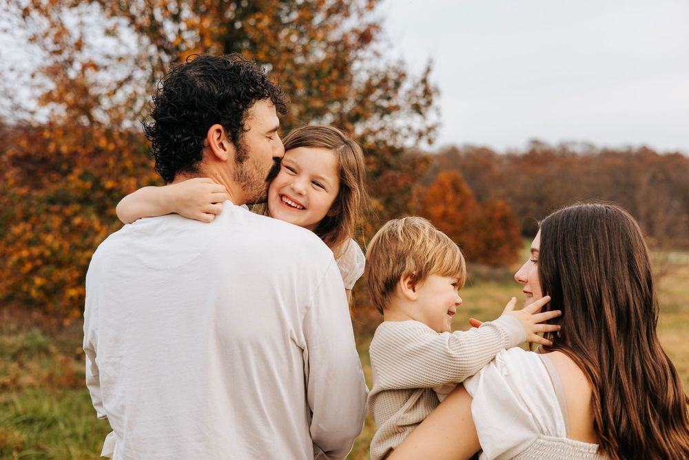 Family of four sharing a joyful moment outdoors in autumn, parents holding children among fall foliage.
