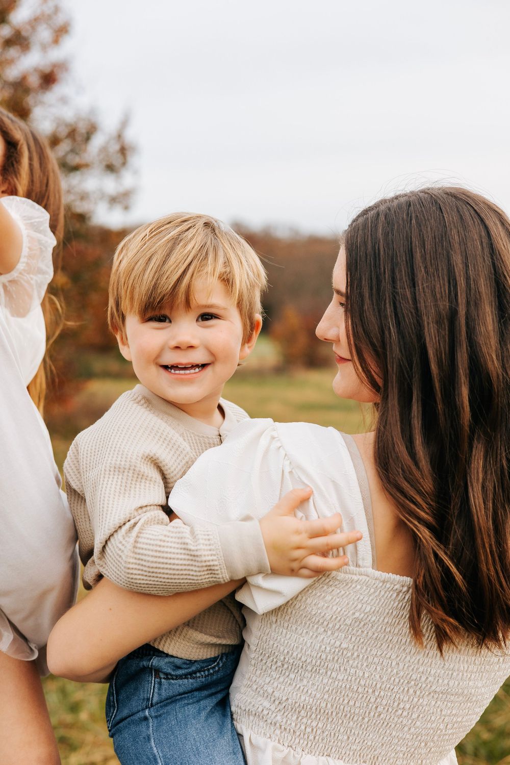 A smiling toddler boy in a beige sweater being held by a dark-haired woman outdoors in an autumn setting.