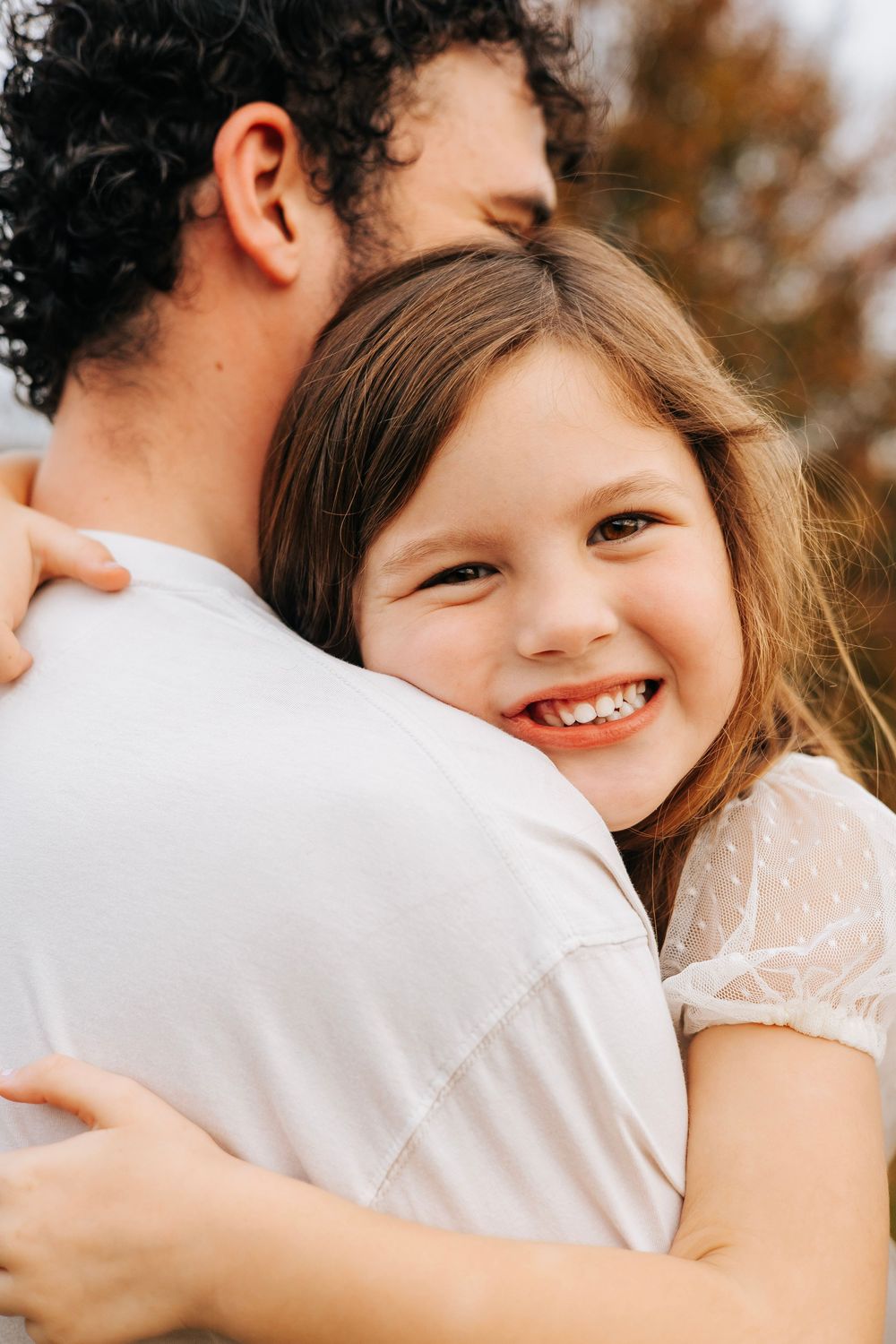 A young girl smiles joyfully while hugging a man in a white shirt outdoors in autumn.