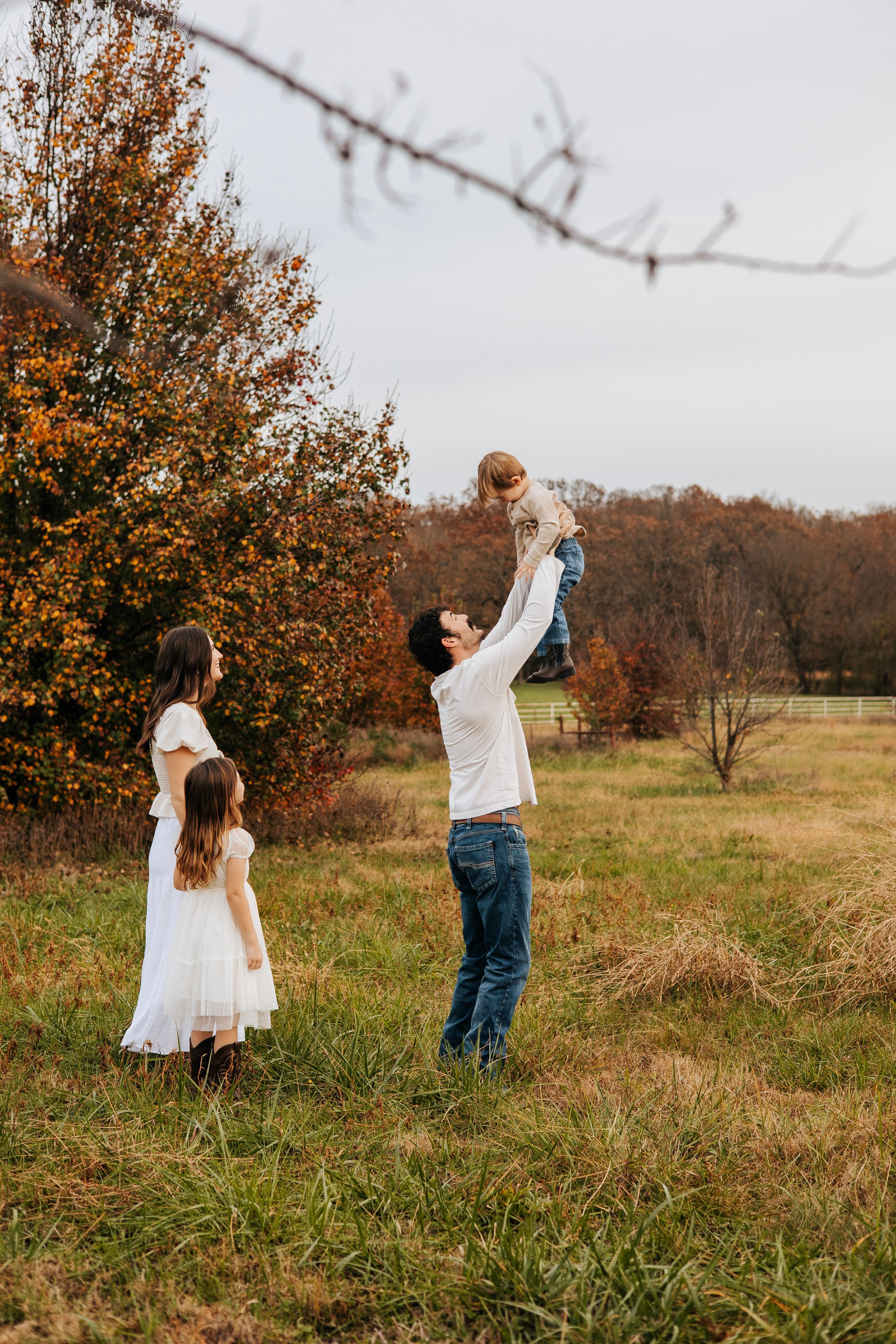 Father joyfully lifts baby in autumn field while mother and daughter watch in matching white dresses.