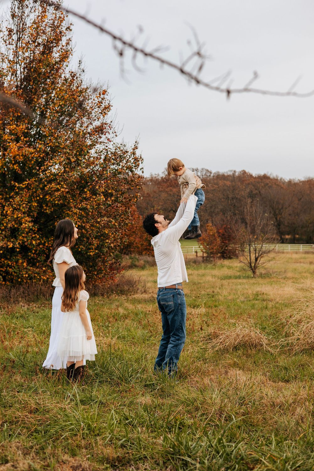 Father joyfully lifts baby in autumn field while mother and daughter watch in matching white dresses.