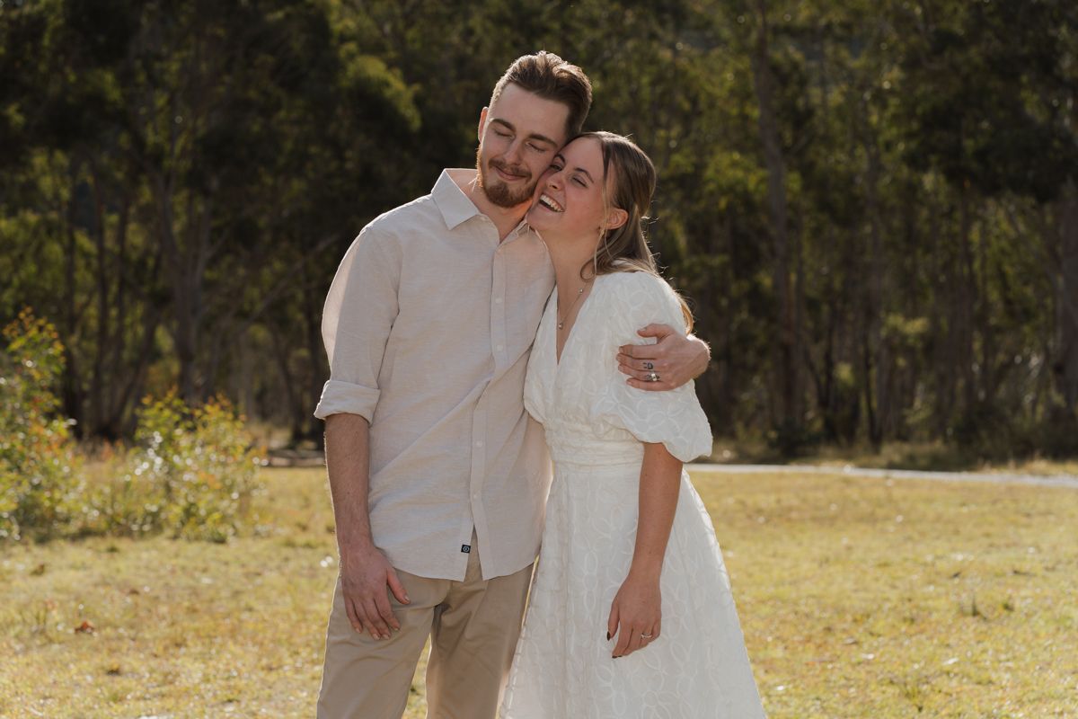 Couple in outdoors sunny field, woman in white dress kissing mans cheek, trees in background during engagement photoshoot