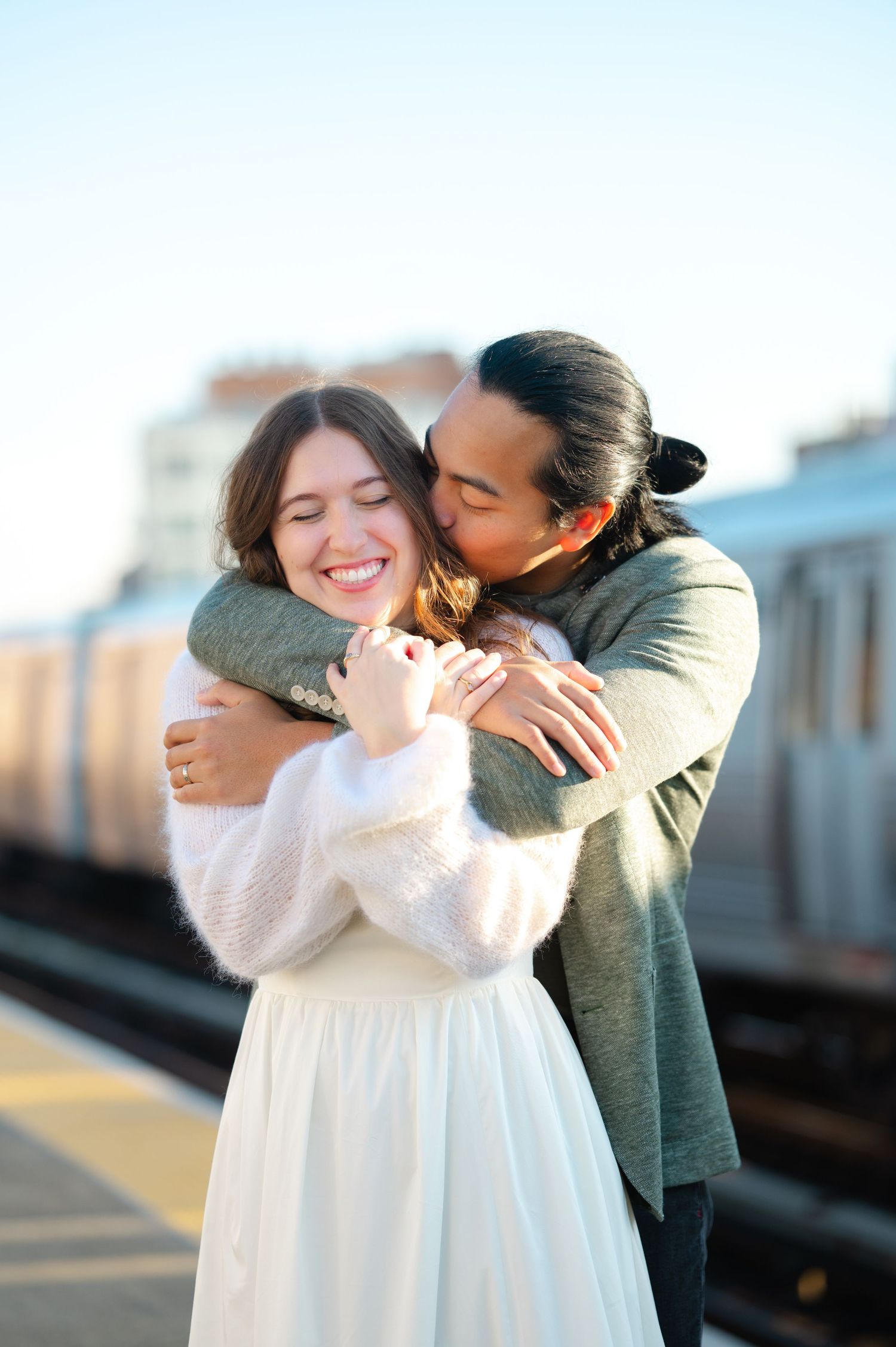 A happy couple embraces on a train platform, the woman smiling as her partner kisses her cheek during a wedding portrait session in Astoria.
