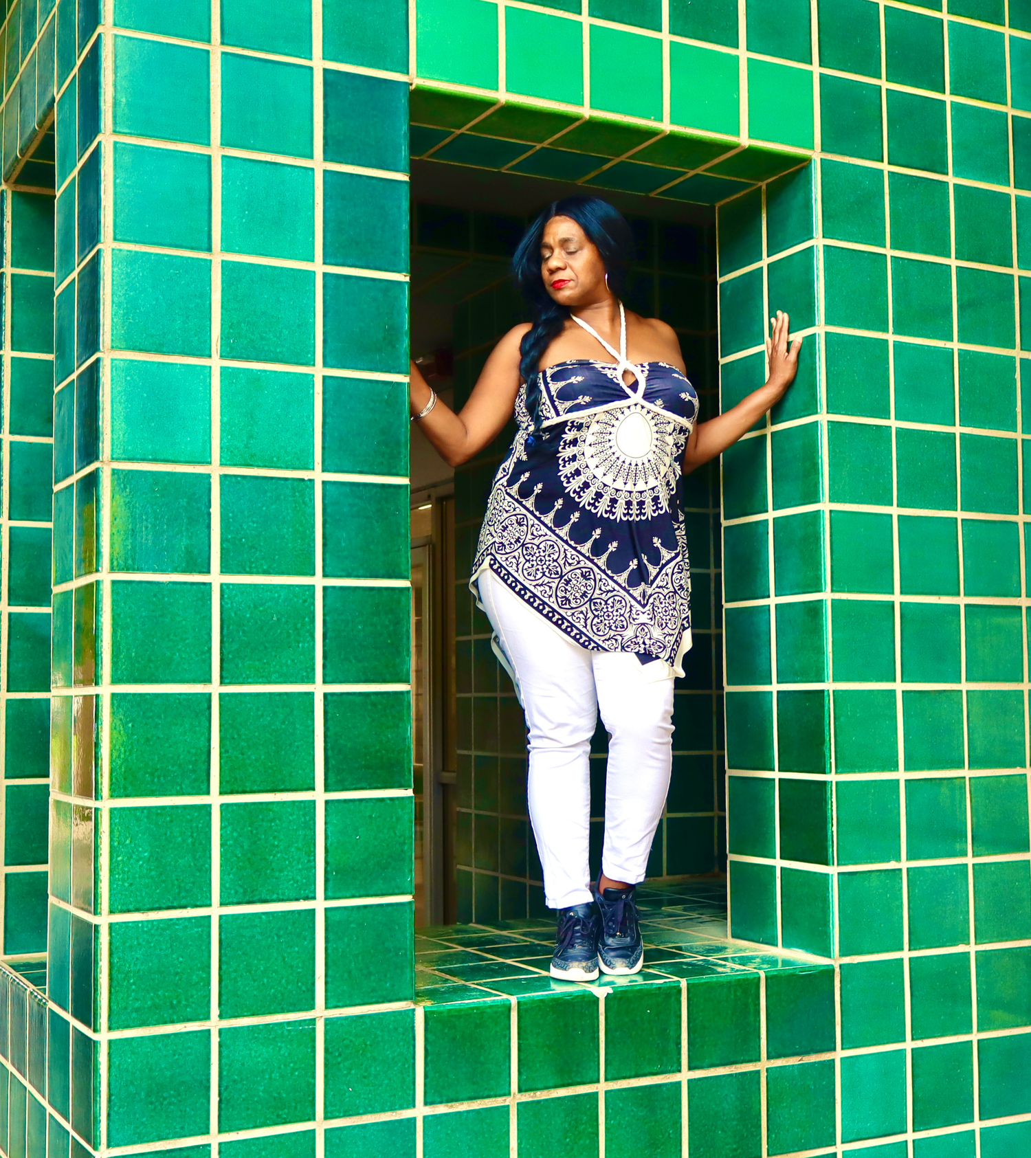 Woman in black and white patterned top and white jeans poses in a green tiled doorway.