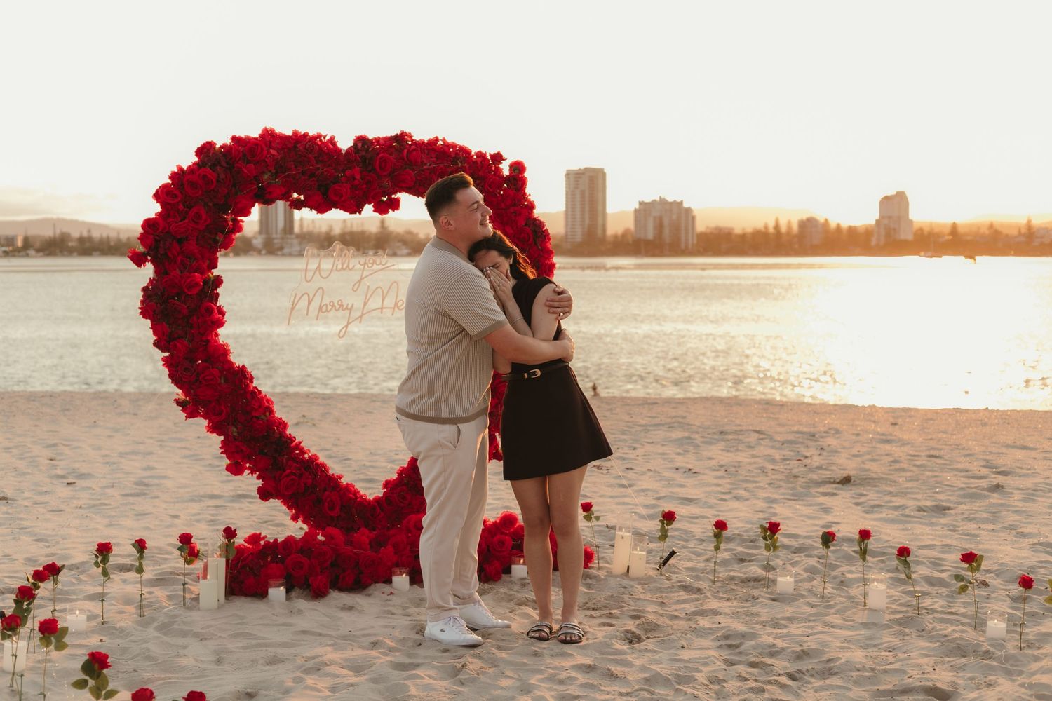 Couple embracing in front of a red rose heart arch on a sandy beach at sunset with city skyline backdrop.