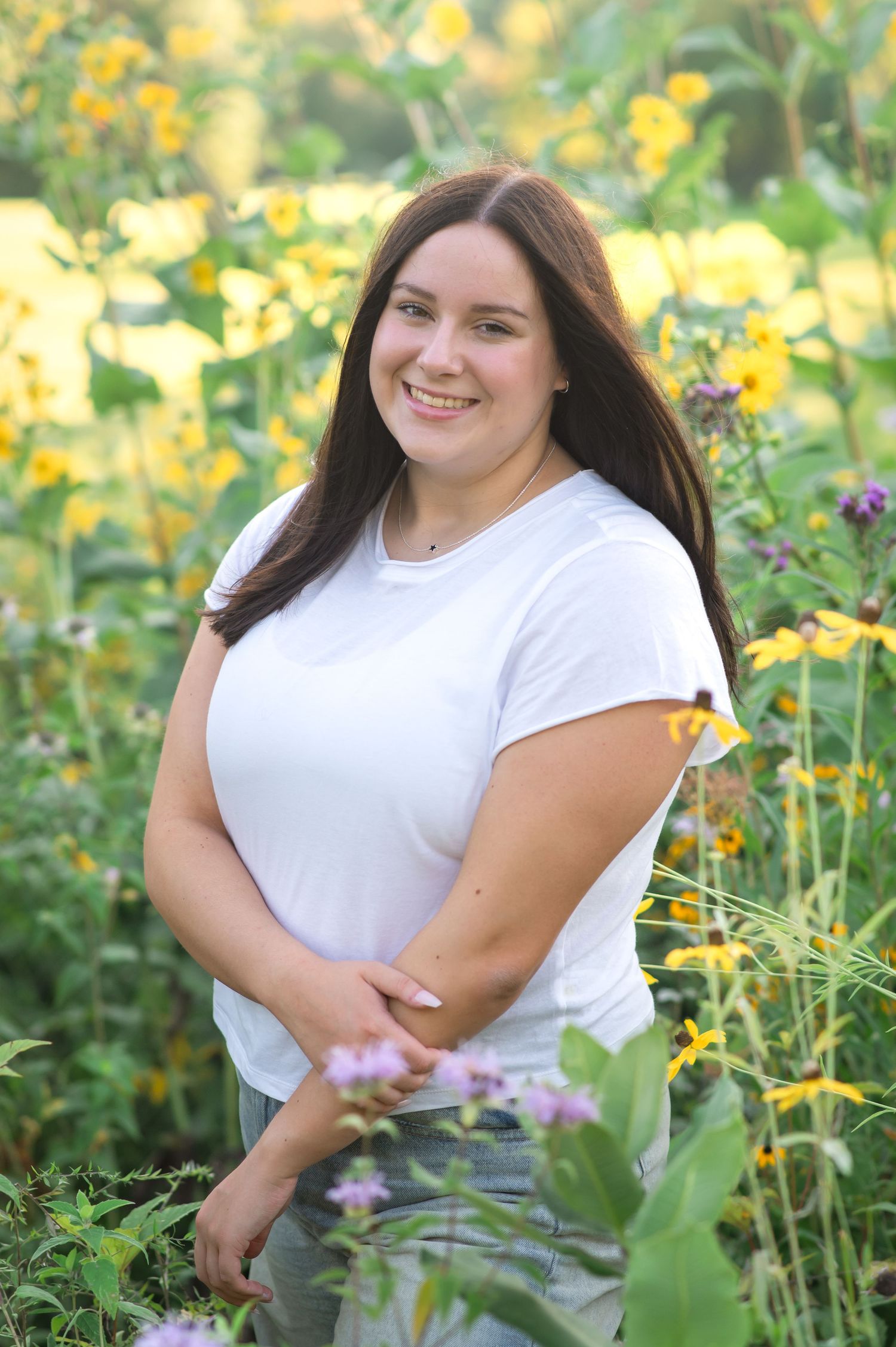 Woman in white shirt poses in garden with yellow and purple wildflowers on sunny summer day.