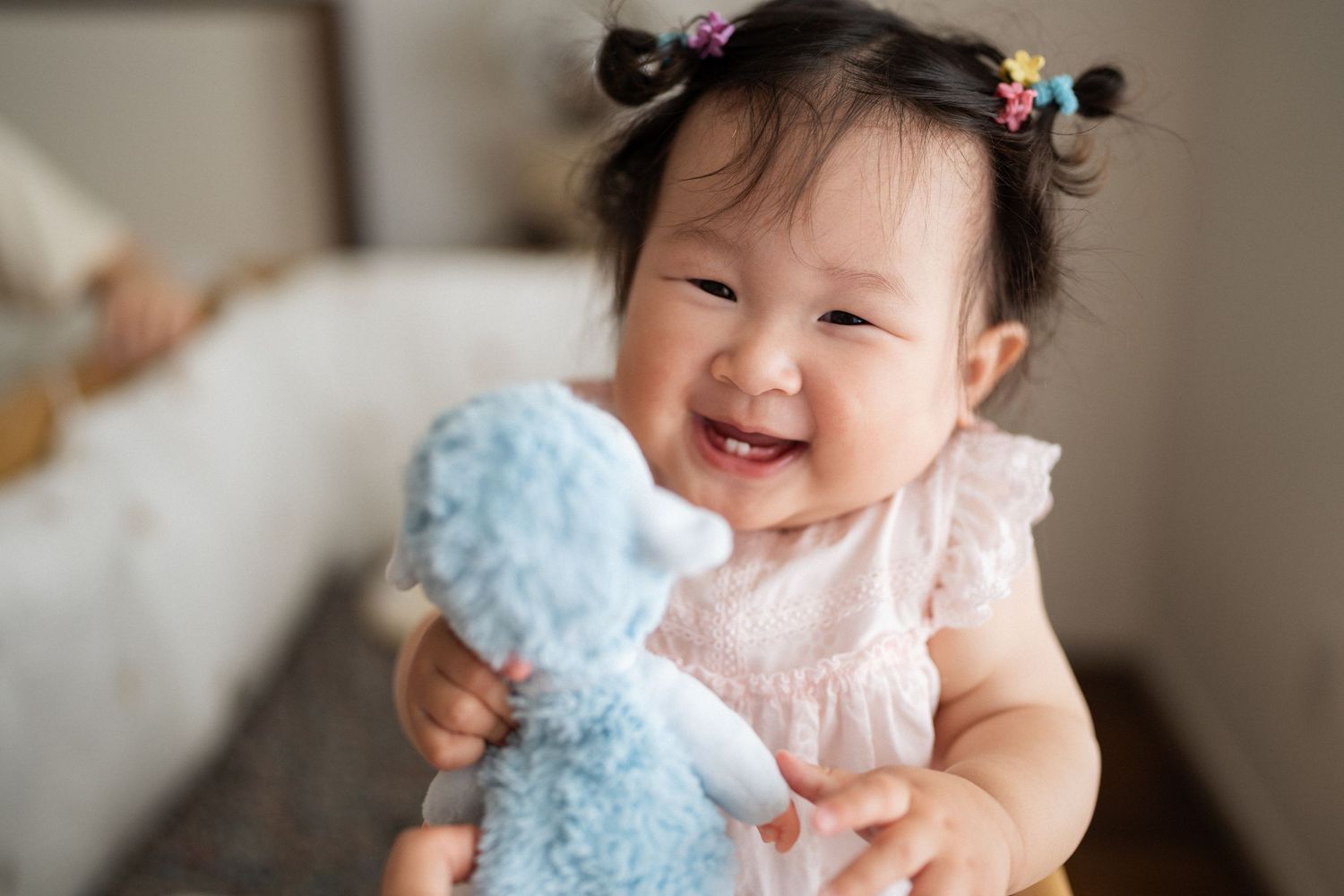 Baby plays joyfully with a soft blue plush toy while wearing a white dress.