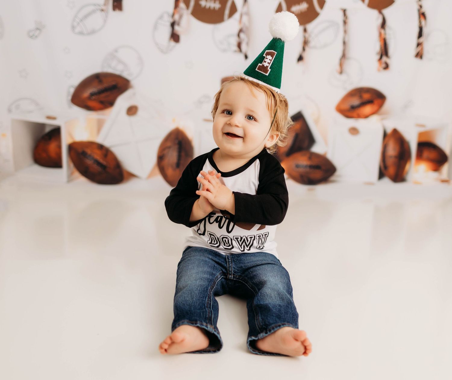 Small child wearing a party hat sits barefoot against white background with football decorations.